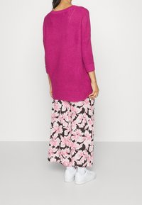 Kvinna klädd i en ljusrosa stickad tröja, blommiga vida byxor med rosa och vita blommor, vita strumpor och vita sneakers, som står med ryggen mot kameran.