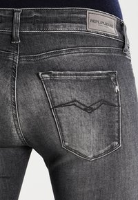 Jeans de mezclilla gris oscuro con una textura desgastada, que presentan un diseño estilizado en el bolsillo trasero y una etiqueta de la marca en la cintura.