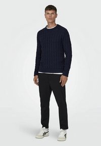 Mornarsko moder pulover z vzorcem kitke, izdelan iz teksturirane tkanine, z okroglim izrezom in rebrastimi manšetami ter spodnjim robom. Združen s črnimi hlačami in belimi supergami.