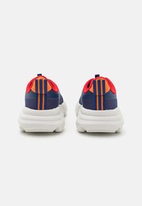Blauwe en rode sportschoenen met een bovenwerk van mesh, felle oranje accenten en dikke witte zolen; heeft verticale strepen op de hiel.