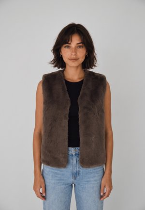 Donna con capelli corti e scuri che indossa un gilet di pelliccia marrone senza maniche sopra una maglietta nera e jeans azzurri, in piedi contro uno sfondo grigio uniforme.