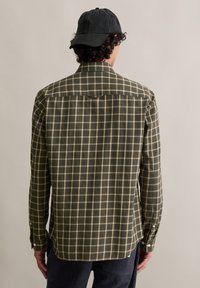 Man met een groen geruit overhemd met lange mouwen, donker corduroy broek en een zwarte baseballcap, met zijn rug naar de camera op een effen beige achtergrond.