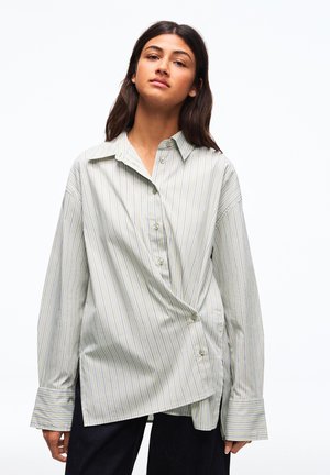 Mladá žena stojaca pred bielym pozadím, oblečená v nadrozmernej svetlošedej pruhovanej košeli na gombíky a čiernych nohaviciach.