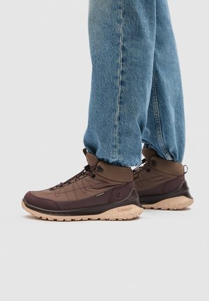 Persona che indossa scarponi da trekking impermeabili marroni con suole beige e jeans blu, in piedi su uno sfondo chiaro.