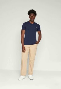 Young man standing with one hand in pocket, wearing a navy blue fitted t-shirt, beige pants, and white sneakers against a plain background.