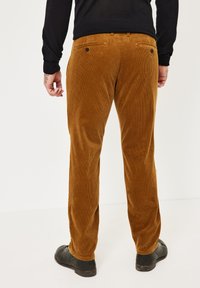 Braune Cord-Hosen mit gerippter Textur, geradem Beinabschluss und zwei Gesäßtaschen mit schwarzen Knöpfen. Getragen mit schwarzen Schuhen.