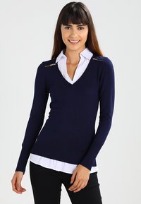 Navy blue sweater with a v-neck, ribbed hem, and two gold zipper accents at the shoulders, layered over a white collared shirt.