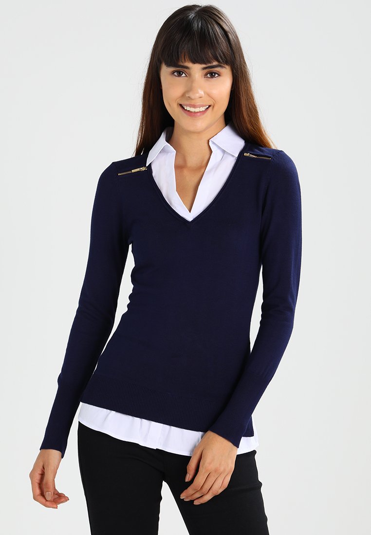 Navy blue sweater with a v-neck, ribbed hem, and two gold zipper accents at the shoulders, layered over a white collared shirt.