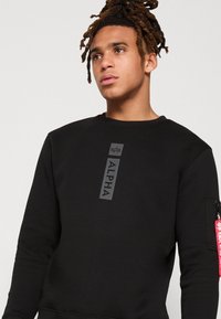 Sudadera negra con un logo en relieve negro "ALPHA" en la parte delantera y un bolsillo con cremallera en la manga con una etiqueta roja. Textura suave.