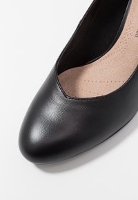 Czarny, fakturowany but na wysokim obcasie z ostrym noskiem i głębokim wycięciem, z wkładką amortyzującą i gładkim wykończeniem wewnątrz.