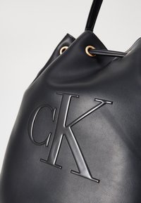 Schwarze Leder-Eimer-Tasche mit geprägtem "CK"-Logo, goldfarbigen Ösen und glatter Textur. Merkmale sind die runde Form und der verstellbare Schulterriemen.