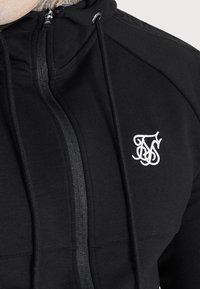 Mustat vetoketjuhupparit, jotka on valmistettu pehmeästä materiaalista. Hupparissa on pieni valkoinen logo rinnassa, hienovaraisia ompelutauksia ja kiristettävä huppu.