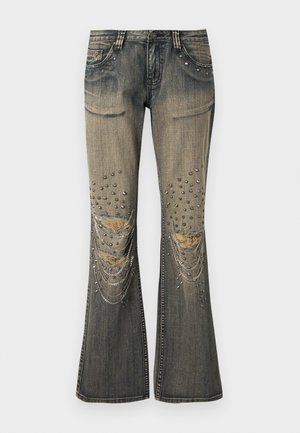 Blugi evazați gri decolorați, cu rupturi uzate la genunchi, capse metalice și ornamente din lanțuri suprapuse pe picioarele din față.