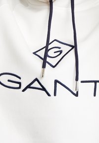 Sudadera blanca con el logo bordado de "GANT" en azul marino y un diseño de diamante. Cuenta con cordones en azul marino con puntas metálicas. Textura suave.
