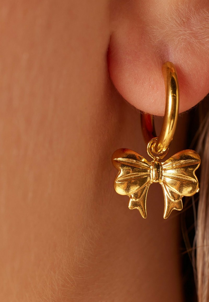 Gouden oorbel met een strikvormige charme. De strik heeft gladde, glanzende oppervlakken en kenmerkende gevouwen randen, wat het ontwerp versterkt.