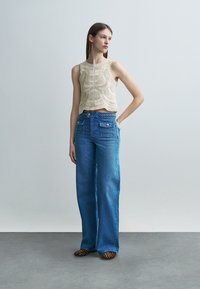 Woman standing, wearing a sleeveless beige crochet top, high-waisted wide-leg blue jeans with front pockets, and leopard print shoes.