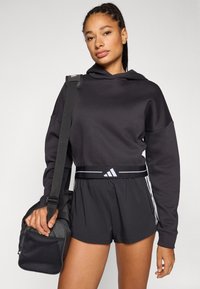 Zwarte hoodie met een ruim ontwerp, gecombineerd met zwarte shorts. Adidas-logo op de tailleband; model houdt een zwarte sporttas met gaasaccenten vast.