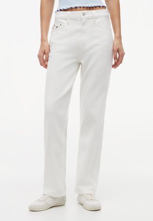 Biele džínsy s rovnými nohavicami na osobe, ktorá má na sebe biele tenisky a krátky svetlomodrý top s volánovým lemom.