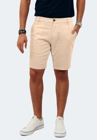 Beige Baumwollshorts mit regulärer Passform, mittelhohem Bund, Fronttaschen und detaillierten Nähten. Kombiniert mit weißen Sneakers.