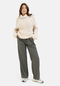 Flauschiger, cremefarbener Strickpullover mit hohem Kragen und geribbelter Textur, kombiniert mit lockeren grauen Hosen und weißen Sneakers mit silbernen Akzenten.