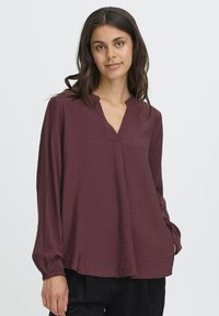 Femme aux longs cheveux noirs portant un blouse maroon ample à manches longues avec un col fendu et un pantalon noir, sur un fond uni.