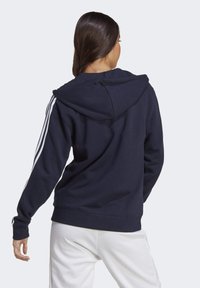 Kvinna med långt mörkt hår som bär en marinblå hoodie med vita ränder på ärmarna och vita byxor, vänd bort mot en enkel bakgrund.