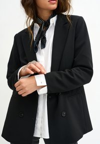 Persona che sistema il polsino di una camicia bianca sotto un blazer nero, indossando una sciarpa a righe bianche e nere su uno sfondo semplice.