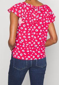 Rote Bluse mit floralen Mustern und kurzen Rüschensleeves, mit einer Schleifendetail auf der Rückseite. Das Material wirkt leicht und hat kleine weiße Blumenmuster.