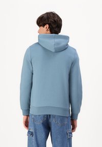 Haut à capuche bleu clair avec une texture douce, il présente une capuche avec cordon de serrage, des poignets et un ourlet côtelés, associé à un jean cargo bleu clair.