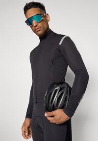 Costume de cyclisme noir avec un col haut, doté d'une poche zippée et d'un accent réfléchissant. La personne tient un casque noir et porte des lunettes de soleil miroir.
