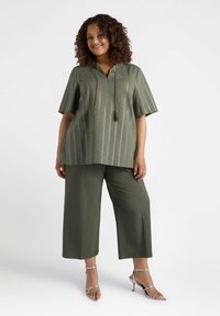 Top de color verde oliva, de manga corta, con rayas verticales y detalles de borlas, combinado con pantalones de pierna ancha en verde oliva. Modelo llevando tacones plateados.