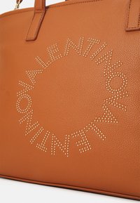 Brun läder toteväska med präglad text omgiven av gyllene nitar, med en strukturerad yta och hållbara handtag.