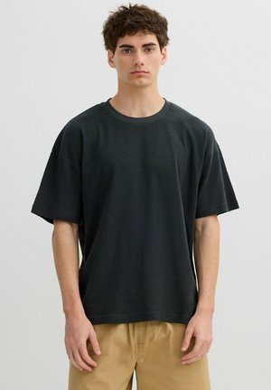 Joven de pie de frente, vistiendo una camisa negra de manga corta y suelta y pantalones beige, sobre un fondo blanco liso.