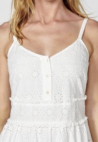 Vit klänning med blommig öljettbroderi, tunna band och knappslå fram. Har en volangmidja och en flödande kjol. Mjuk tygtextur.