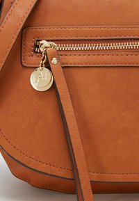 Detale torebki z brązowej skóry z złotym zamkiem błyskawicznym i okrągłym złotym zawieszką z wygrawerowanym napisem "A1" na uchwycie zamka.