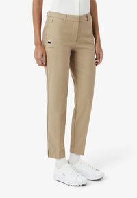 Pantaloni beige con vestibilità sartoriale, dotati di una tasca frontale e un piccolo logo verde. Abbinati a scarpe da ginnastica bianche e calzini bianchi.