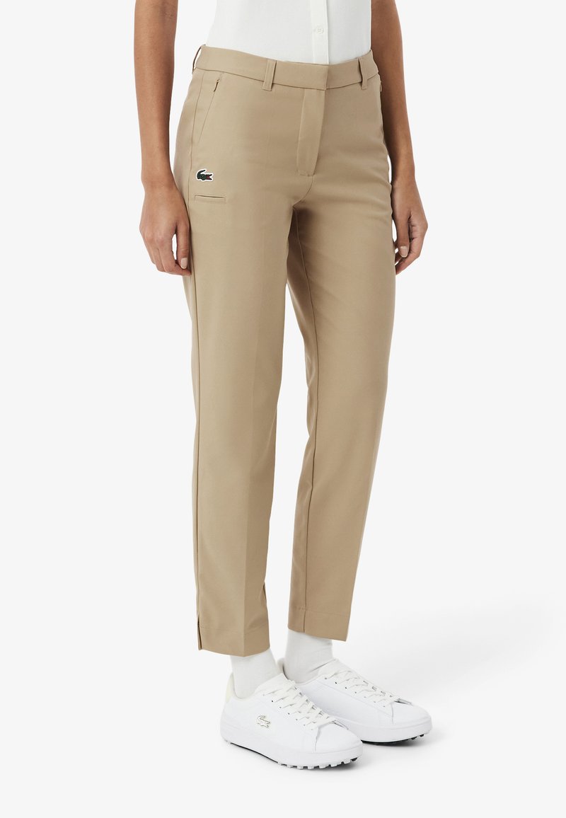 Pantaloni beige con vestibilità sartoriale, dotati di una tasca frontale e un piccolo logo verde. Abbinati a scarpe da ginnastica bianche e calzini bianchi.