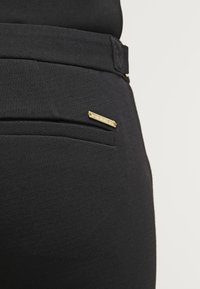 Pantalons noirs au toucher lisse, dotés d'une poche arrière et d'un petit accent en métal doré. Design ajusté avec une coupe élancée.