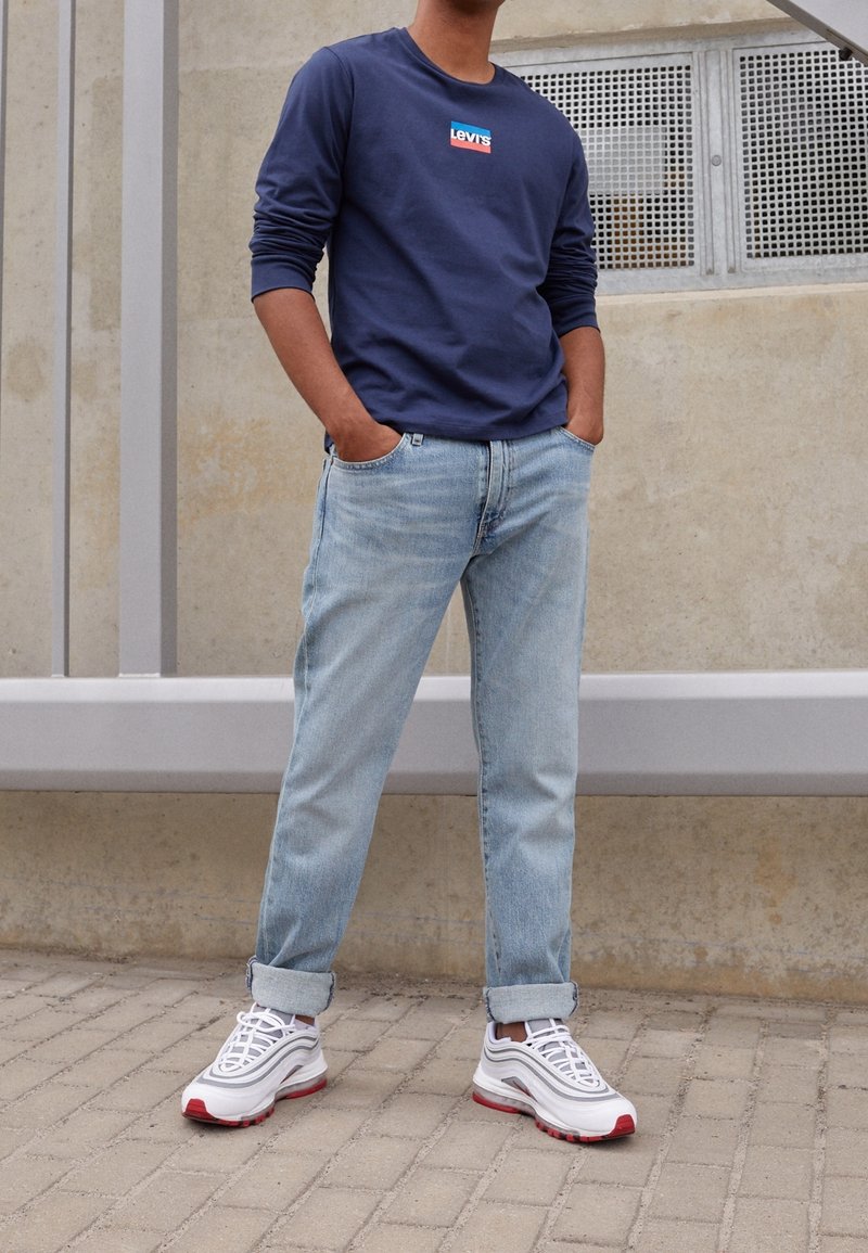 Henkilö seisoo kädet taskuissa, päällään tummansininen Levi's-sweatshirt, käärittyjä vaaleansinisiä farkkuja ja valkoisia lenkkitossuja, joissa on punaiset pohjat.