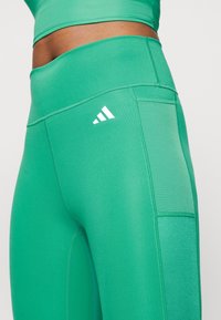 Zielone legginsy sportowe z elastycznego materiału, z szerokim pasem w talii, przeszyciami i małym białym logo Adidas z boku.