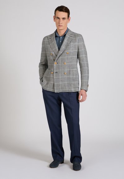 Hombre con blazer gris de cuadros de doble botonadura, camisa azul, pantalones azul marino y zapatos oscuros, de pie con una mano en el bolsillo contra un fondo gris.