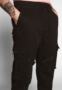 Black cargo pants with elastic waistband, drawstring, and multiple zippered pockets. The fabric is smooth with a slightly matte finish.