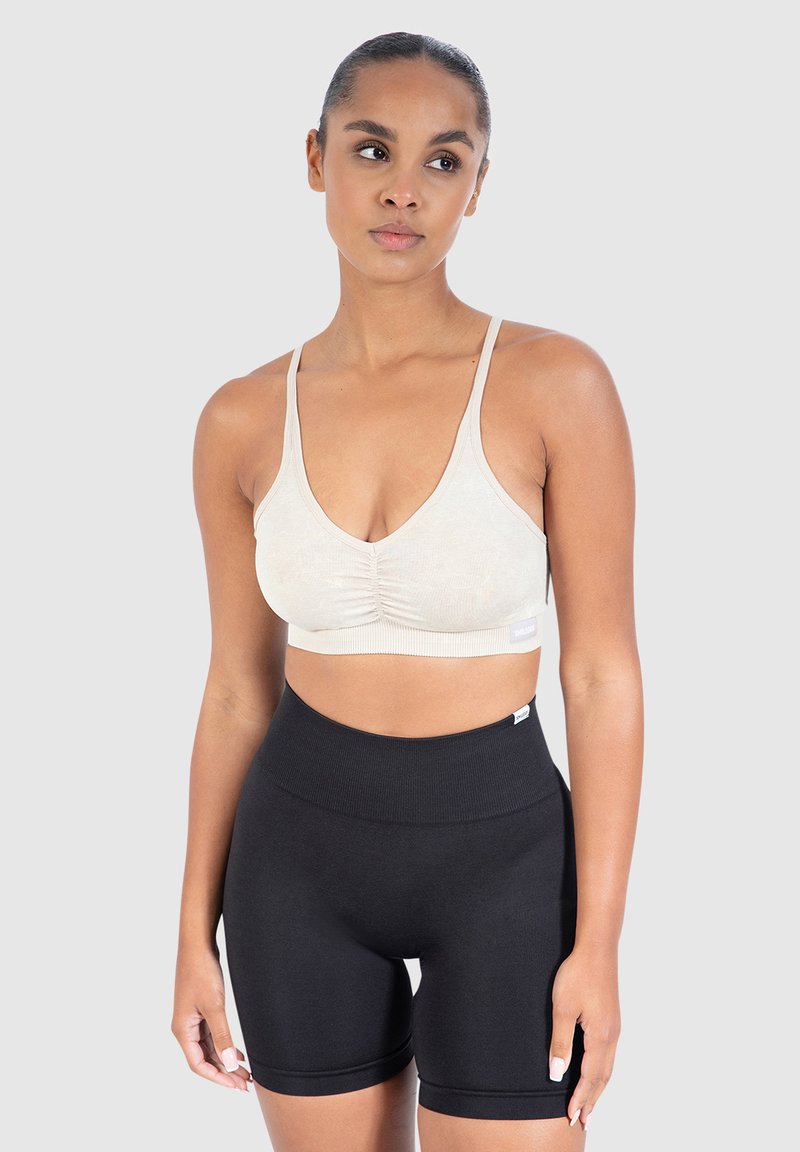 Urheiluliivi vaaleanbeige, jossa on tekstuuripinta ja poimutettu etuosa; yhdistetty korkeavyötäröisten mustien shortsien kanssa. Molemmat ovat joustavia ja myötäileviä.