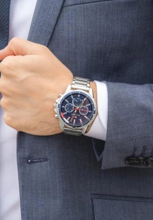 Muškarac u sivom odijelu podešava srebrni metalni ručni sat s tamnoplavim brojčanikom i više podbrojčanika na lijevom zapešću.