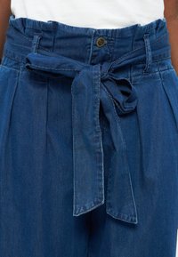 Μπλε τζιν παντελόνι με ψηλή μέση, υφασμάτινη ζώνη δεμένη σε φιόγκο, κλείσιμο με κουμπί και θηλιές για ζώνη, φορεμένο με λευκό τοπ.