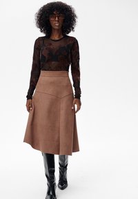 Kobieta z kręconymi włosami, ubrana w brązową, kwiatową bluzkę z przezroczystego materiału, beżową spódnicę w kształcie litery A, czarne wysokie kozaki oraz oversize'owe brązowe okulary przeciwsłoneczne.