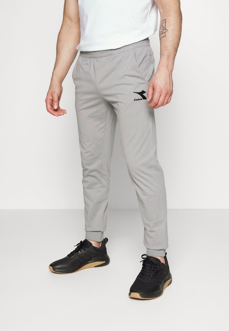 Man die lichtgrijze joggerbroek met zwart logo en zwarte sneakers draagt en tegen een witte achtergrond staat.