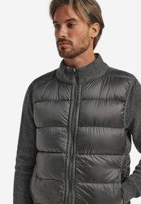 Grau gepolsterte Weste mit hohem Kragen aus glänzendem Material, übereinander getragen über einem dunkelgrauen Langarmshirt. Vorderreißverschluss und Seitentaschen sichtbar.