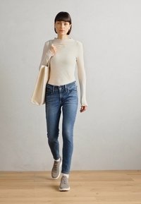 Nő, aki bordázott, krém színű, hosszú ujjú felsőt és világoskék skinny farmert visel, bézs táska a kezében, szürke sportcipőben, fapadlón.