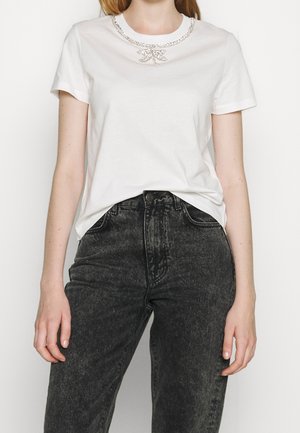 Kobieta w białym T-shircie z wzorem naszyjnika z cyrkoniami i czarnych, wysokich jeansów z efektem acid wash, stojąca na jednolitym tle.